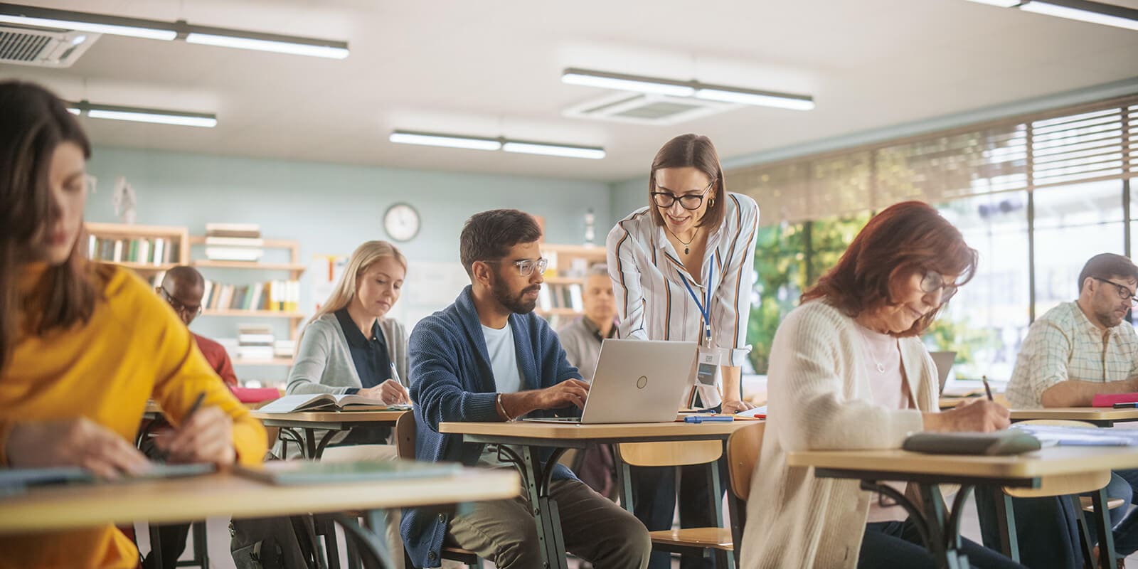 A teacher in classroom of students helping one student with their laptop