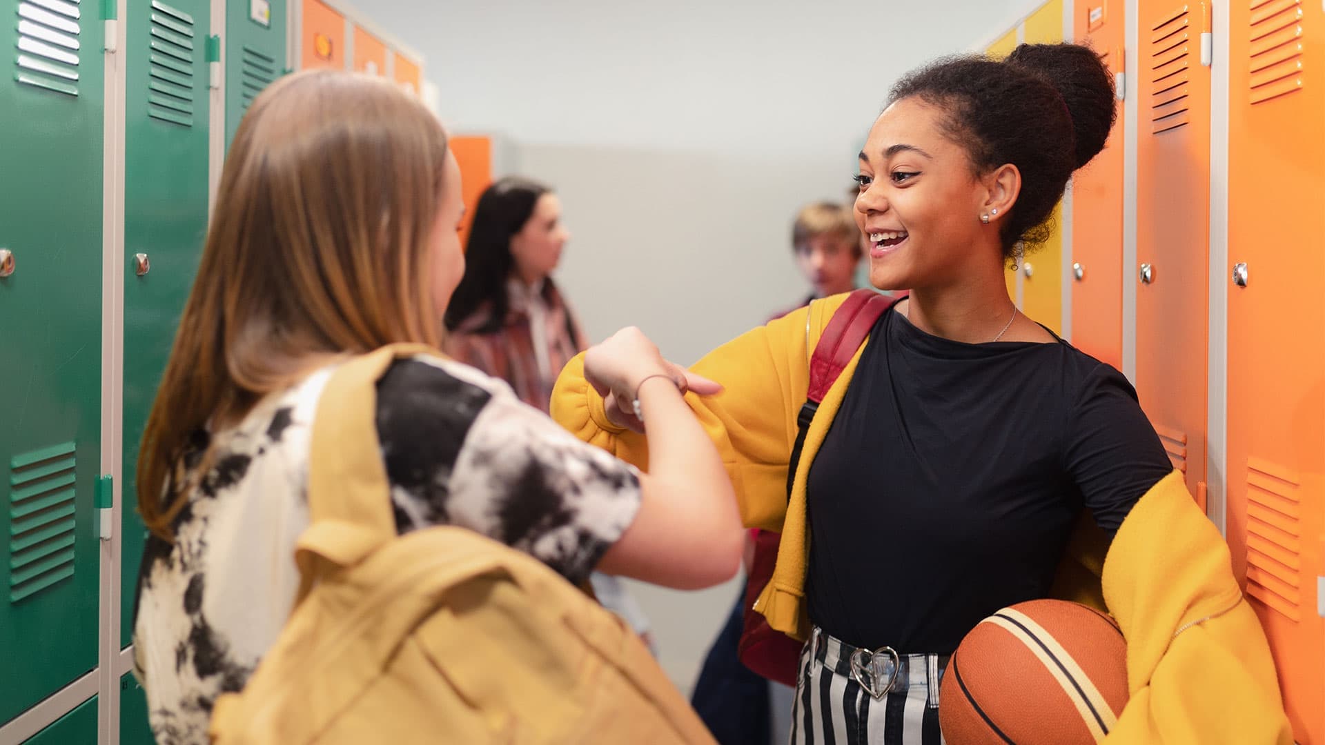 Two students interact in a school hallway, surrounded by colorful lockers, one holding a basketball and wearing a bright yellow cardigan.