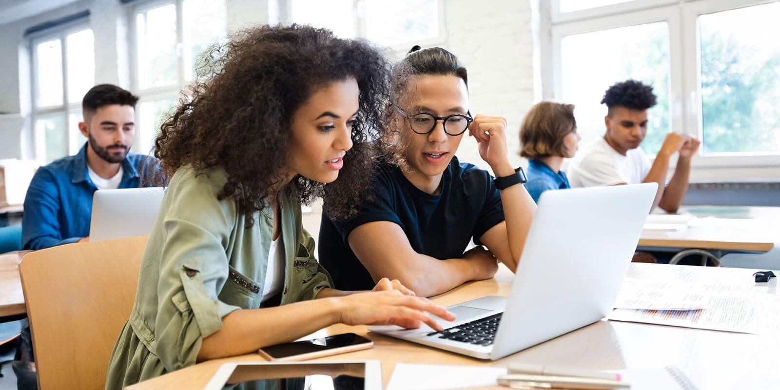Students in a classroom setting, using laptops and engaging in discussion, with large windows providing natural light in the background.