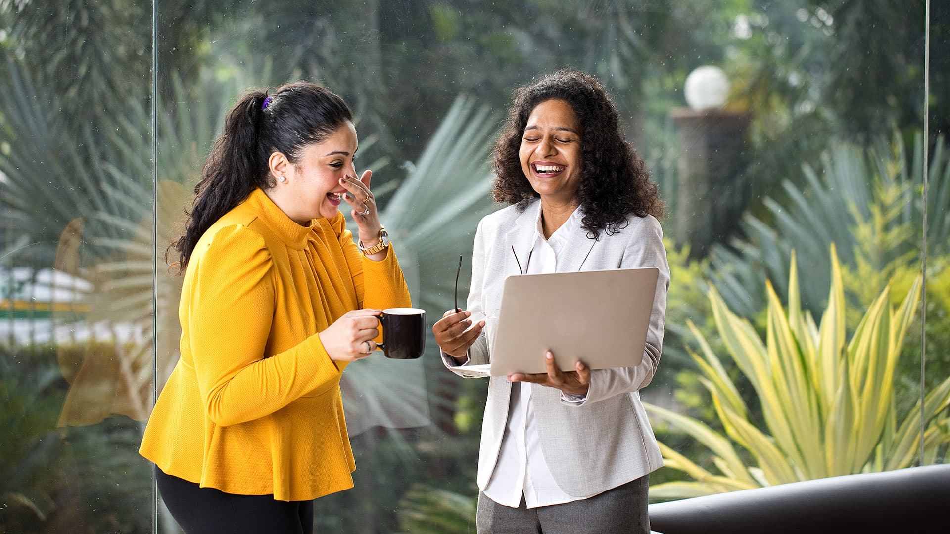 Two women standing outdoors, laughing while holding a laptop, with trees and plants in the background.